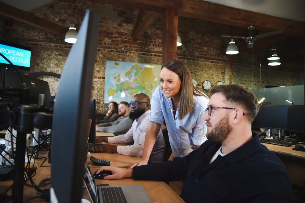Two people, a man and an woman, are looking at the same monitor in an open-space office
